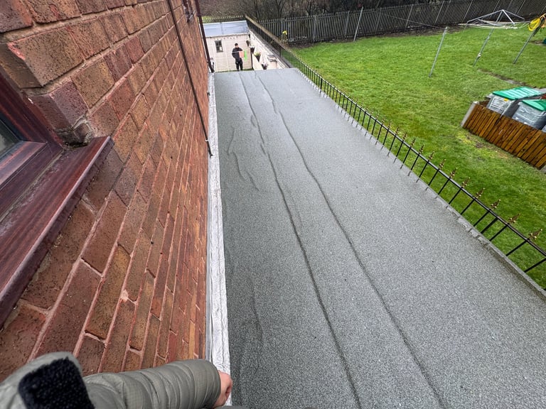 Overhead view of a brick building wall, paved walkway with lane markings, green lawn with fencing, and small figures in the distance