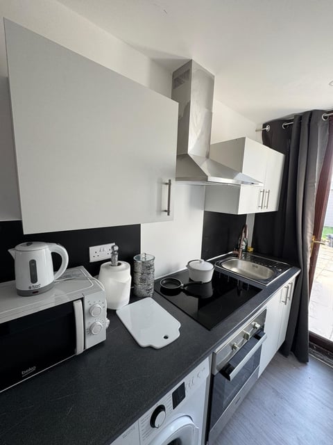 Compact kitchen with white and black cabinetry, electric cooktop, kettle, microwave, and window with black curtains