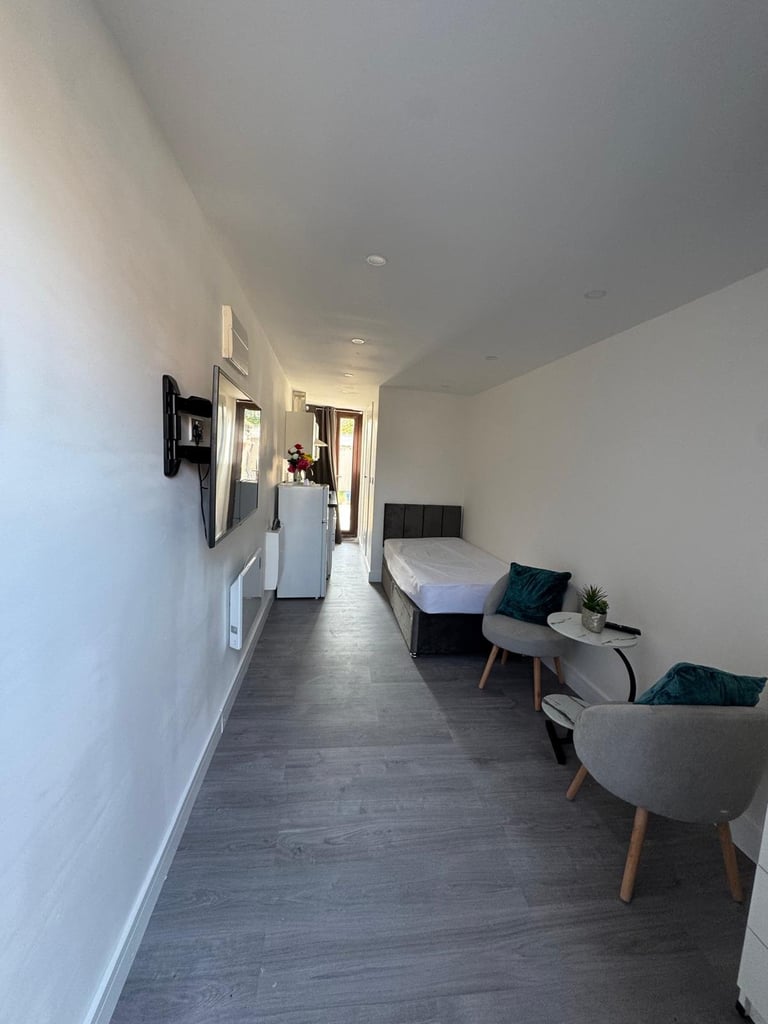 Modern studio apartment interior with white walls, dark hardwood flooring, mounted TV, bed, and seating area along a hallway leading to a kitchen
