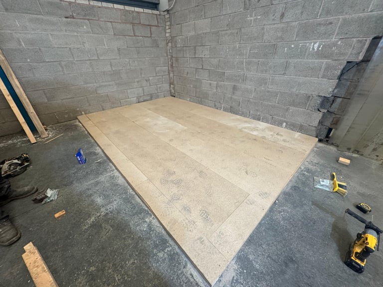 Large plywood sheet installed on concrete floor in basement with gray brick walls and construction tools scattered around