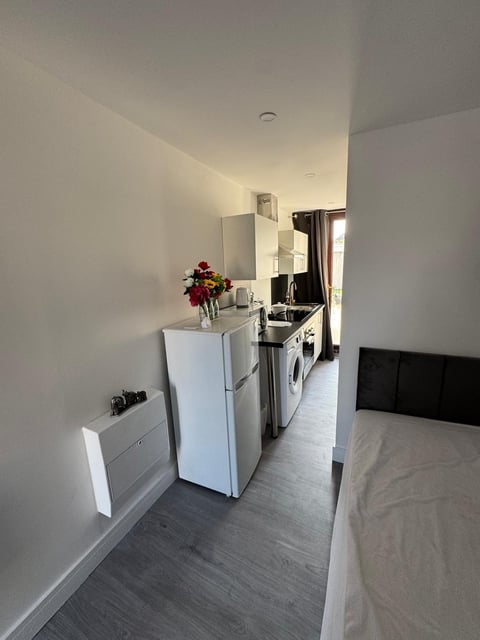 Modern studio apartment kitchen area with white appliances, flowers on counter, and bed visible on right