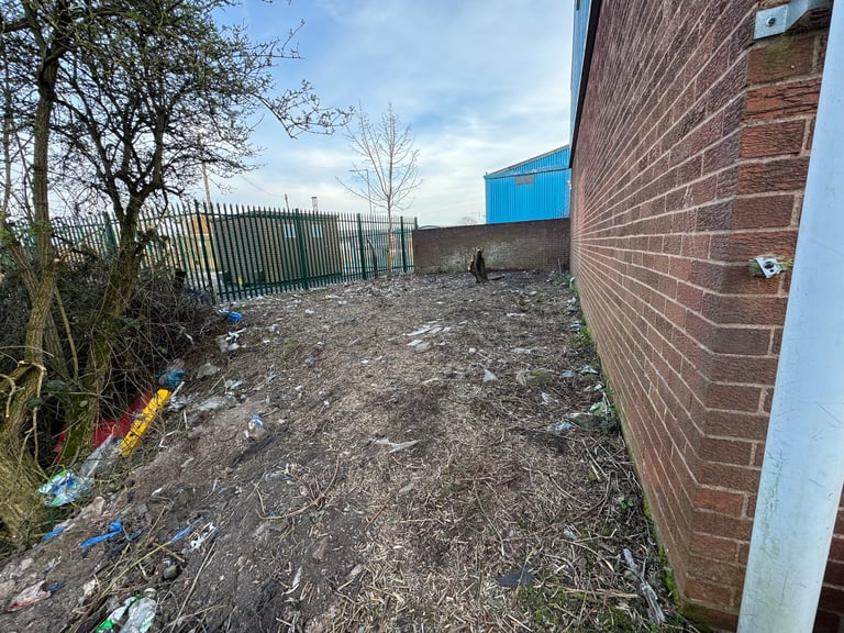 Overgrown outdoor yard area with brick building, metal fence, and debris scattered on bare ground
